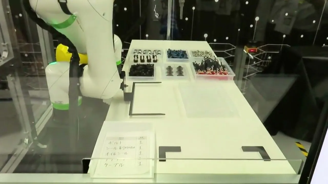 A white FANUC CRX cobot at a demo bench with trays of bolts and seals and a handwritten parts list in Japanese in the foreground